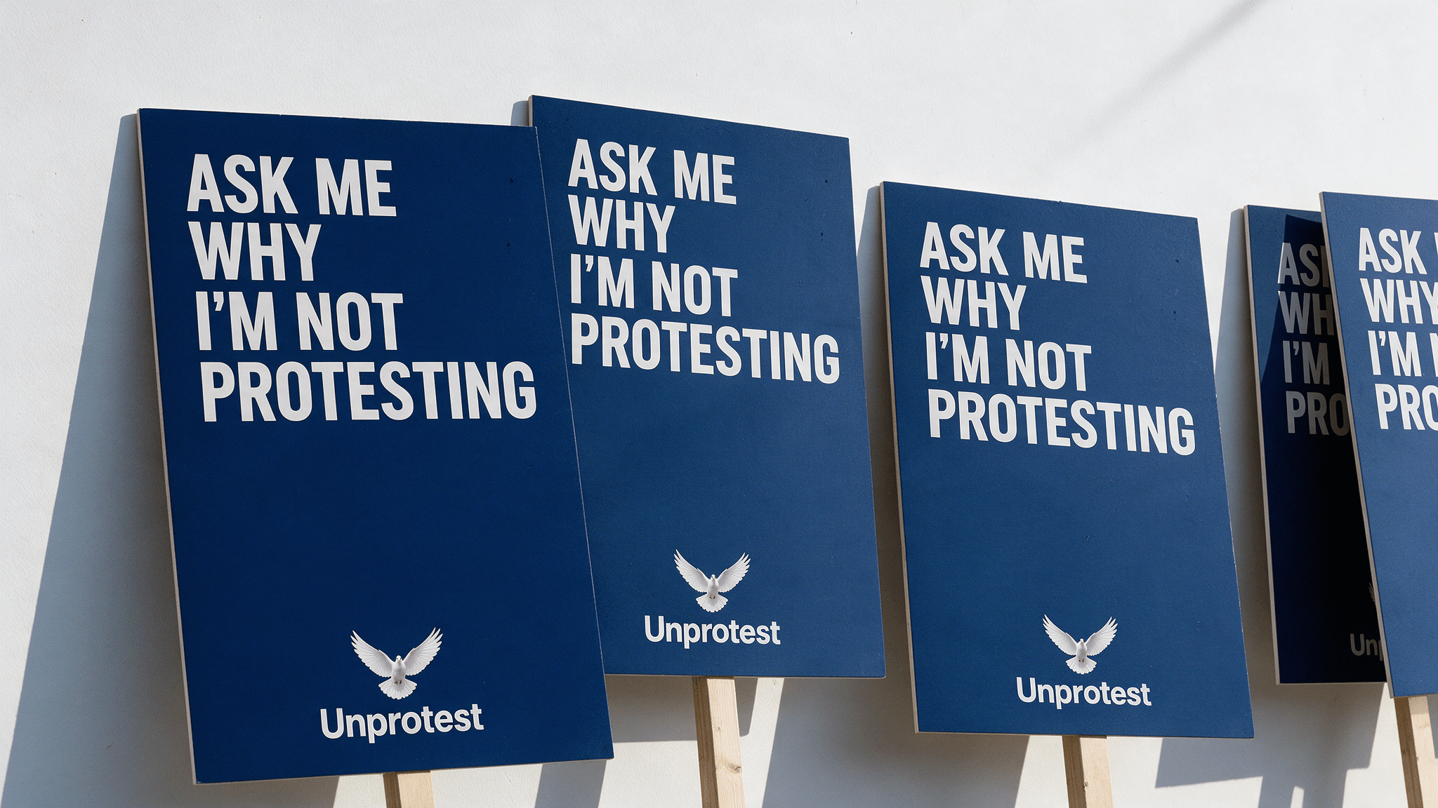 Blue Unprotest placards lined up against a white wall, each reading: Ask me why I’m not protesting.